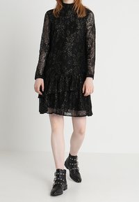 Abito in pizzo nero con maniche lunghe e orlo a balze svasato. Abbinato a stivaletti neri con accenti in metallo sulla fibbia.