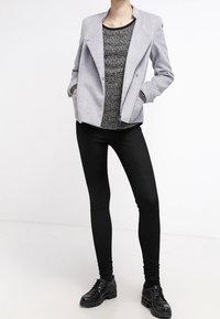 Blazer gris tejido con cuello de muesca y cierre de doble botón, llevado sobre una blusa negra con estampado, combinado con jeans skinny negros y zapatos.