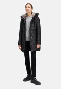 Kobieta w czarnej pikowanej zimowej kurtce z kapturem wykończonym futrem, w szarym swetrze, czarnych spodniach i masywnych czarnych butach, stojąca na jednolitym tle.