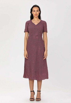 Femme debout portant une robe midi à manches courtes rouge et noire à motifs, avec des talons noirs à bride à la cheville, sur un fond uni.