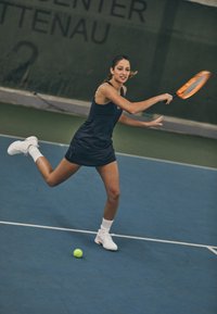 Jugador de tenis vestido con un vestido azul marino y zapatillas blancas, preparado para golpear una pelota amarilla en una pista azul con un fondo verde.