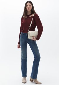 Burgunderfarbener Strickpullover mit Ausschnittdetails; blaue Jeans mit leichten Gebrauchsspuren; beigefarbene Handtasche mit gestepptem Muster; beigefarbene High Heels.