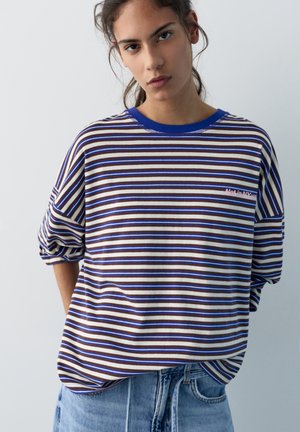 Mladá žena s vážnym výrazom tváre, oblečená v pruhovanom tričku s dlhým rukávom v hnedej, bielej a modrej farbe a v svetlomodrých džínsoch, stojaca na jednoduchom pozadí.