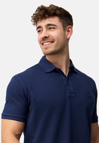 Marineblå polo skjorte laget av teksturert bomullsstoff, med en to-knapps glidelås og en liten logo på brystet. Korte ermer.