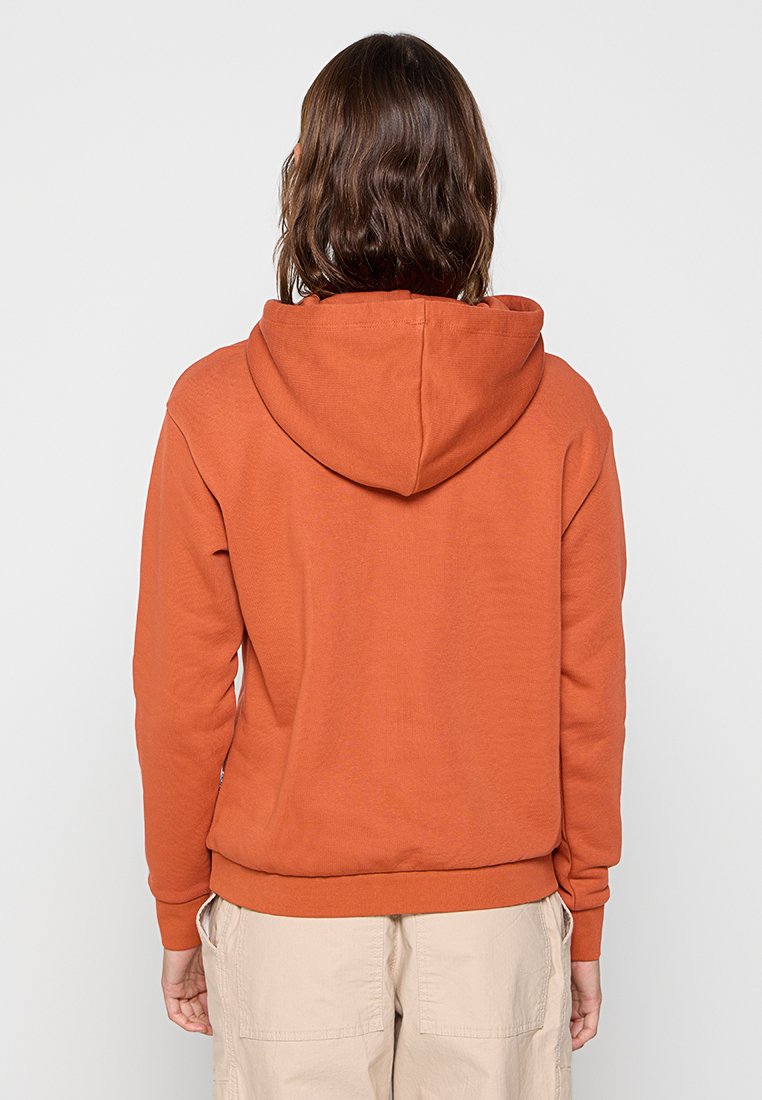 Osoba z średniej długości brązowymi włosami, ubrana w pomarańczową bluzę z kapturem i beżowe spodnie, stoi tyłem na białym tle.