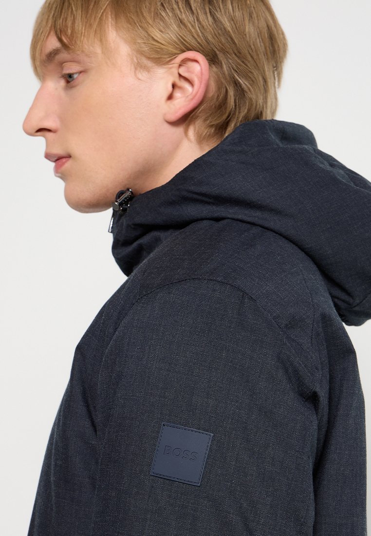 Veste à capuche bleu marine foncé en tissu texturé, dotée d'un logo distinctif sur l'épaule et d'une fermeture éclair au niveau du cou.