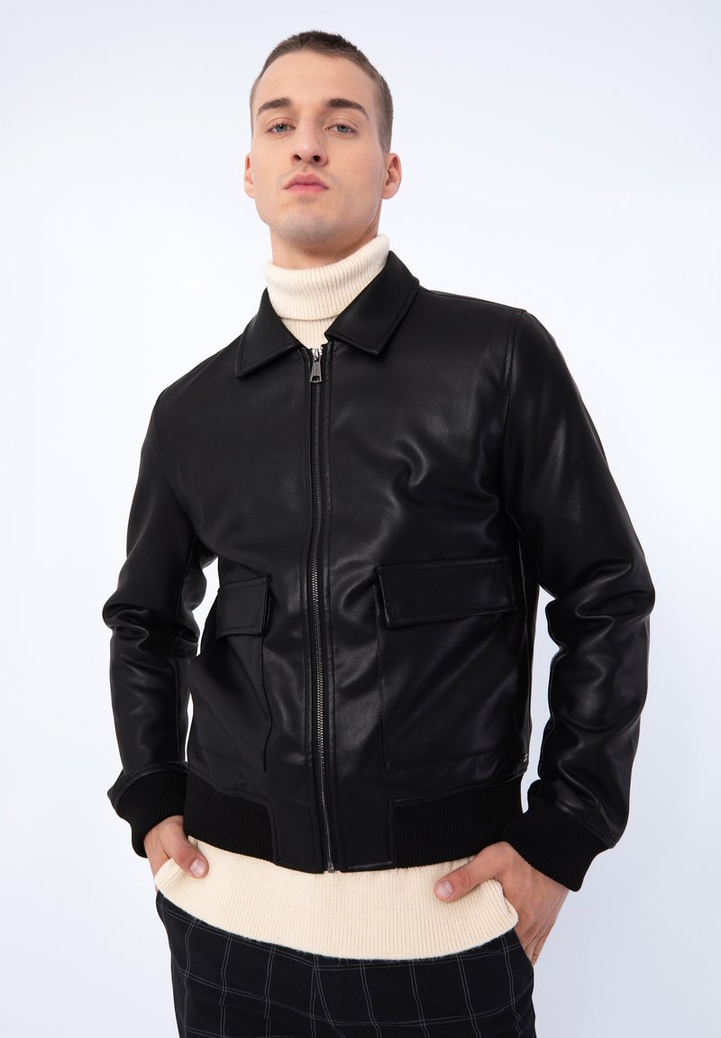 Veste en cuir noir avec un col, zip avant, deux poches poitrine, poignets et ourlet côtelés, associée à un col roulé beige. Texture lisse.