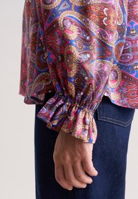 Langärmlige Bluse mit lebhaftem Paisleymuster in lila, rot und goldenen Farbtönen. Verfügt über geraffte Manschetten und eine fließende Silhouette.