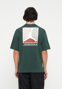 Tamsiai žalios spalvos medvilninė marškinėlė su dideliu logotipu ant nugaros, kuriame pavaizduota stilizuota figūra ir po ja užrašas "JORDAN" baltomis raidėmis. Trumpomis rankovėmis, laisvas kirpimas.
