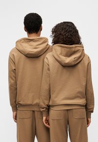 Καφέ βαμβακερές φούτερ μπλούζες με κουκούλα και ριμπ μανσέτες, φαίνονται από πίσω. Συνδυασμένες με καφέ παντελόνια με τσέπες φακέλους.