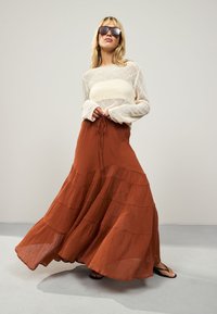 Lengvas kreminės spalvos megztinis su ilgomis rankovėmis ir rūdžių spalvos sluoksniuota maxi sijonas. Sijonas turi raištelį juosmens srityje ir lengvą, krentančią medžiagą.