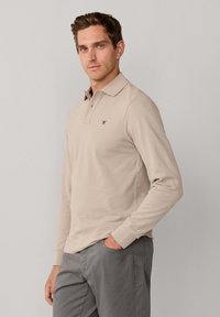 Polo beige à manches longues avec col et logo bleu marine ; texture douce et coupe ajustée, assorti à un pantalon gris.