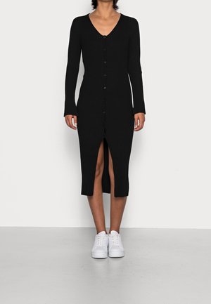 Vrouw draagt een zwarte geribde midi-jurk met knopen, lange mouwen en een voorste split, gecombineerd met witte sneakers, staand tegen een effen achtergrond.