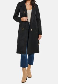 Schwarzer doppelreihiger Trenchcoat mit goldenen Akzenten, großem Kragen und langen Ärmeln. Getragen über einem beigen Oberteil und blauen, ausgestellten Jeans.