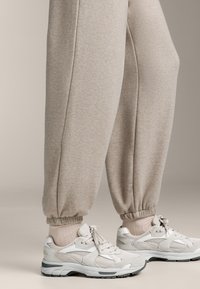 Beige joggingbroek met elastische boorden, gecombineerd met witte en grijze sneakers. Sneakers hebben een mix van mesh en suède materialen met vetersluiting.