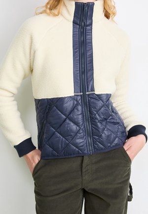 Persona que lleva una chaqueta de forro polar beige con tejido acolchado azul marino en la mitad inferior y cremallera delante, manos en los bolsillos de pantalones verde oscuro.