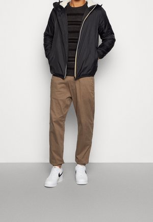 Man wearing black hooded jacket with white fleece lining, black striped sweater, beige pants, and white sneakers with black swoosh logo.