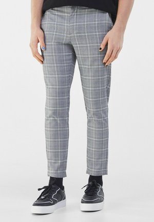 Personne portant un pantalon gris à carreaux, des chaussettes noires et des baskets noires à semelles blanches, debout les mains le long du corps.