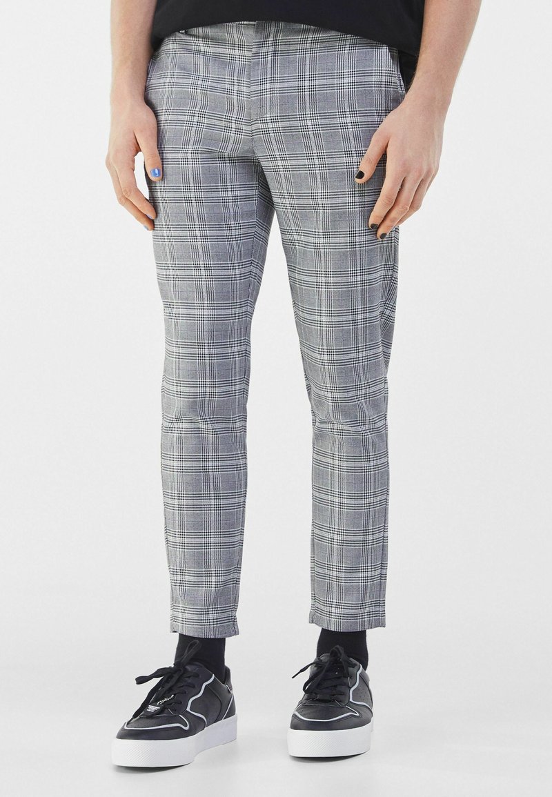 Personne portant un pantalon gris à carreaux, des chaussettes noires et des baskets noires à semelles blanches, debout les mains le long du corps.