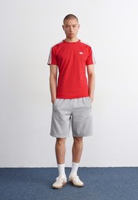 Raudona trumpa rankovė T-maikė su baltomis juostelėmis ir logotipu ant krūtinės, derinama su pilkais šortais ir baltais sportbačiais su raudonais akcentais.