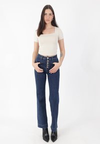 Camiseta ajustada color beige claro con mangas cortas combinada con jeans azul oscuro de talle alto que tienen cierre de cinco botones y un ligero acampanado. Botas negras puntiagudas.