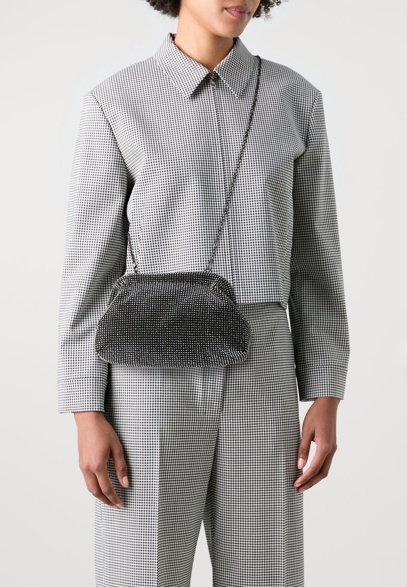 Femme portant une chemise à carreaux noir et blanc à manches longues et un pantalon avec un petit sac bandoulière noir texturé sur l'épaule.