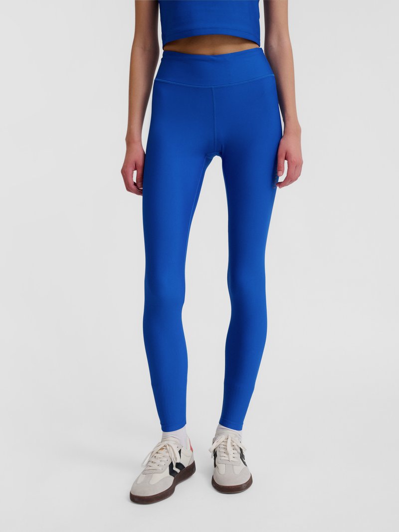 Kvinde iført lyseblå leggings med høj talje, hvide sneakers og en matchende blå cropped top, stående mod en ensfarvet lys baggrund.