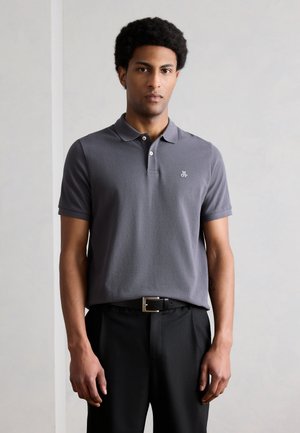 Mężczyzna stojący w pomieszczeniu, ubrany w szarą koszulkę polo z krótkim rękawem, czarne spodnie i czarny pasek, zwrócony przodem, z neutralnym wyrazem twarzy.