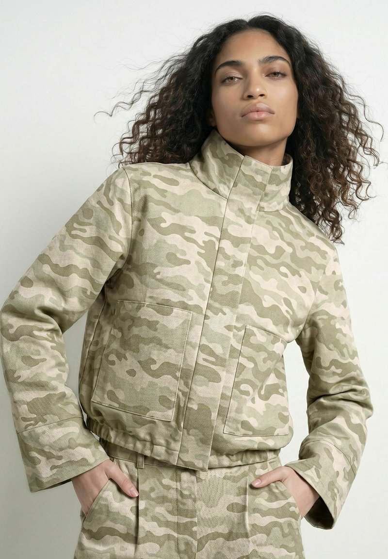 Femme aux cheveux bouclés portant une veste camouflage beige et vert clair ainsi qu'un pantalon assorti, les mains dans les poches, sur un fond uni.