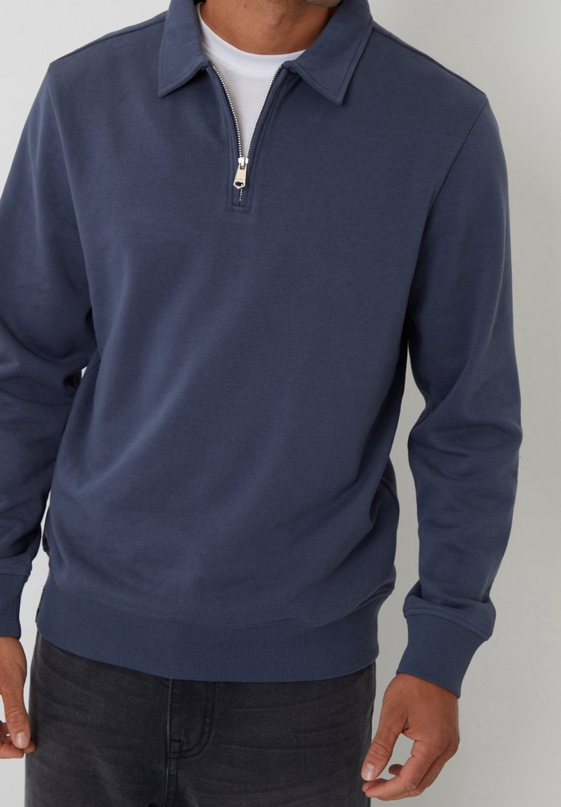 Homme portant un pull bleu marine à manches longues avec une fermeture éclair sur un quart, par-dessus un sous-vêtement blanc et un pantalon foncé, debout devant un fond uni.