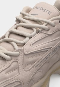 Μπεζ αθλητικό παπούτσι Lacoste με υλικό από καστόρι με υφή, λεπτομερή ραφή και χοντρές στρογγυλεμένες κορδόνια δεμένα μερικώς στο μπροστινό μέρος.