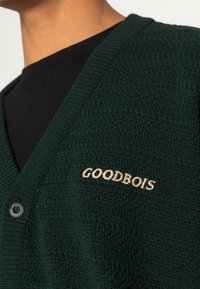 Nahaufnahme einer Person, die einen dunkelgrünen Strickcardigan mit einem beigen, auf der Brust gestickten "GOODBOIS"-Logo trägt.