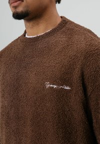 Bruine fleece trui met een gestructureerd oppervlak, voorzien van een ronde hals en geborduurd "Young Atelier" in het wit op de borst.