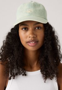 Young woman with curly dark hair wearing a light blue Levi’s cap and a white ribbed tank top, looking directly at the camera.