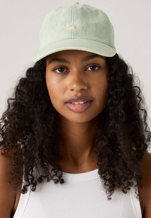 Young woman with curly dark hair wearing a light blue Levi’s cap and a white ribbed tank top, looking directly at the camera.