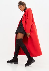 Abrigo de lana rojo sobre un vestido negro, combinado con botas de ante negras hasta la rodilla con un tacón bajo y grueso y detalle de cremallera.