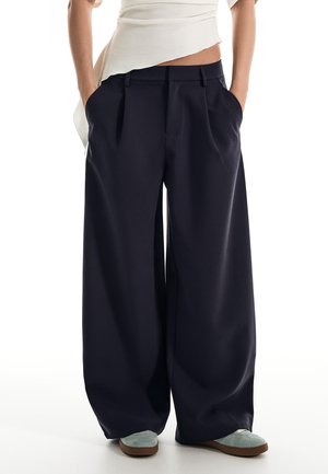 Vista dalla vita in giù di una persona che indossa pantaloni a gamba larga blu scuro, con le mani nelle tasche, abbinati a sneaker azzurro chiaro e una maglietta bianca.