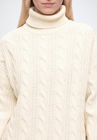 Femme portant un pull à col roulé en maille torsadée couleur crème avec un motif texturé, montré des épaules à la bouche.