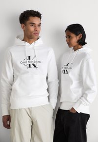 Bijele dukserice s prednjim džepom, crni logo "Calvin Klein Jeans" jasno otisnut na prsima. Obojica modela nose svijetle i tamne hlače.