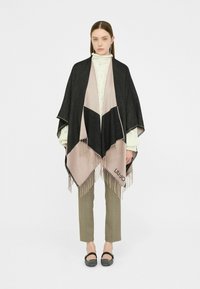 Poncho nero e rosa con bordi frangiati, design triangolare, indossato sopra un dolcevita crema, abbinato a pantaloni verde oliva e ballerine nere.
