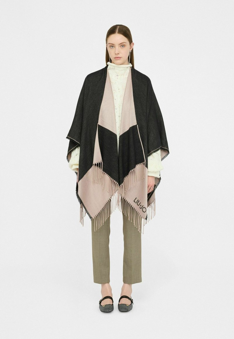 Poncho nero e rosa con bordi frangiati, design triangolare, indossato sopra un dolcevita crema, abbinato a pantaloni verde oliva e ballerine nere.