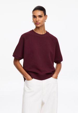 Femme aux cheveux foncés attachés porte un sweat-shirt bordeaux à manches courtes et un pantalon blanc avec les mains dans les poches sur un fond blanc.