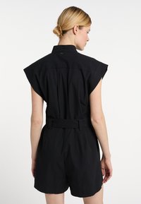 Salopetă neagră din bumbac, cu guler, mâneci scurte și o croială lejeră, cu talie ajustabilă printr-un brâu; textură netedă și detalii de design minime.