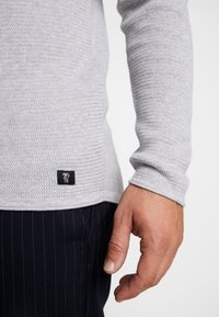 Grå stickad långärmad topp med en strukturerad yta, prydd med en svart etikett med vit branding vid fållen. Bärs över randiga byxor.