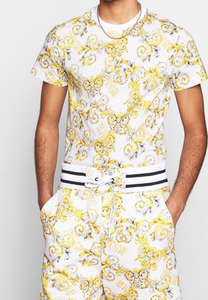 Homme portant une chemise blanche à manches courtes et un short avec un imprimé baroque floral jaune et une taille à cordon rayée noir et blanc.