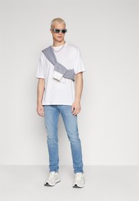 Hvit t-skjorte, lyseblå jeans, grå genser drapert over skulderen, hvite joggesko med svarte detaljer, iført grå solbriller, kort blond hår.