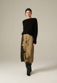 Femme portant un haut noir à manches longues, une jupe midi à motifs marron et des bottes noires jusqu'aux genoux, se tenant devant un fond beige uni.