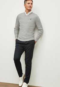 Pull en tricot gris avec un col en V et un logo bleu, associé à un pantalon foncé et des baskets blanches. Texture lisse, coupe décontractée et design moderne.