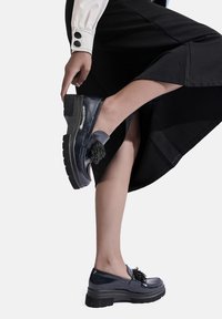Scarpe navy in pelle verniciata con suola spessa, caratterizzate da un accento decorativo con nappina. Il design è elegante con punta tonda e texture liscia.