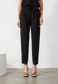 Pantalon noir fuselé avec ceinture à nouer, en tissu lisse. Comprend une taille haute et une longueur chevilles coupées. Associé à des talons à brides.